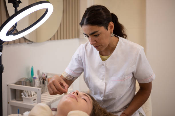 Argentinean young woman during eyebrow lamination and correction procedure. Beautician young woman appliyng cream, tweezing eyebrows and combing them. Buenos Aires - Argentina